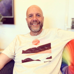 A smiling man with a beard sitting on a couch in a well-lit room, wearing a white T-shirt.