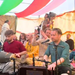 People attending a colorful indoor event with draped fabric and string lights, some playing musical instruments, others listening and taking photos.