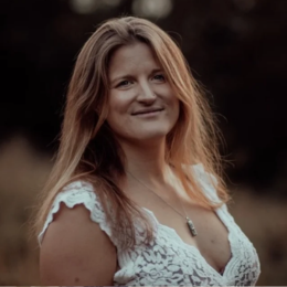A woman with long, wavy hair smiling outdoors during sunset.