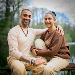 A smiling man and woman sitting outdoors on a bench, embracing each other, with trees and blue sky in the background.