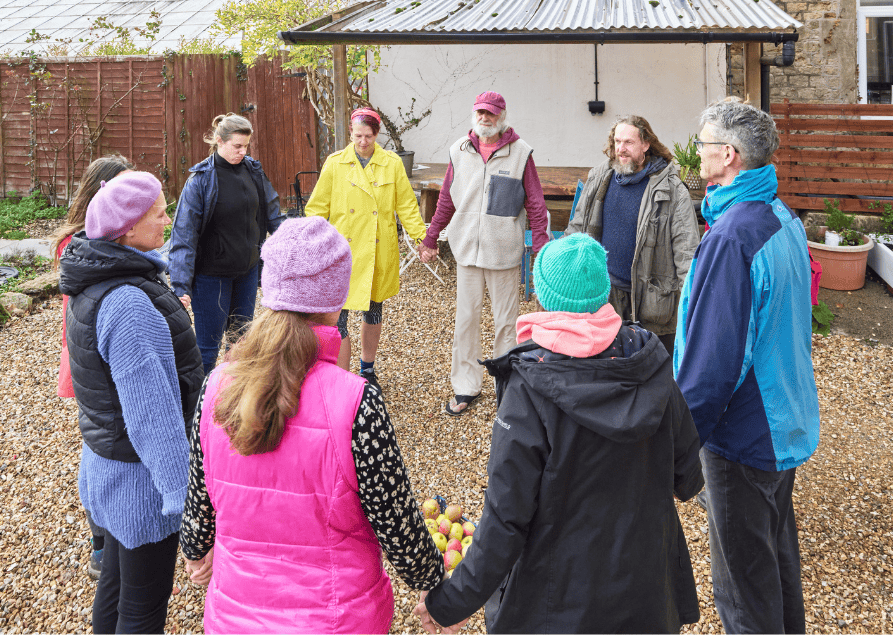 Group of people holding hands in a circle outdoors in a garden on a gravel surface.