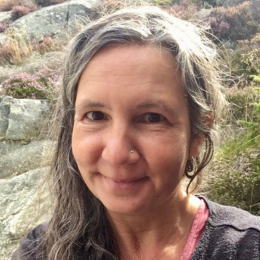 A woman with long gray hair and a nose piercing smiling outdoors with a rocky background and autumn foliage