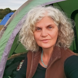A woman with curly gray hair taking a selfie outdoors in front of tents.