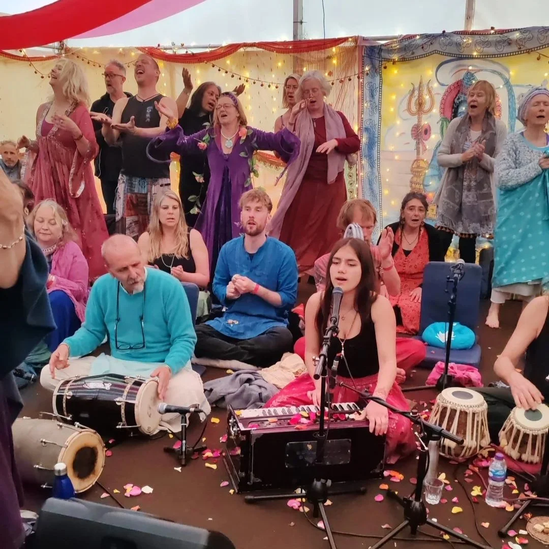 A large group of people, including singers and musicians, participate in a cultural or religious celebration inside a decorated tent, with flowers and string lights. Some are singing with expressions of joy, while others are playing drums and a keyboard. Confetti is scattered on the floor, creating a festive atmosphere.