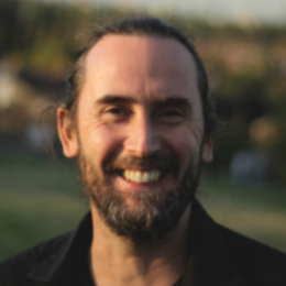 A smiling man with a beard and long hair pulled back, outdoors in a natural setting.
