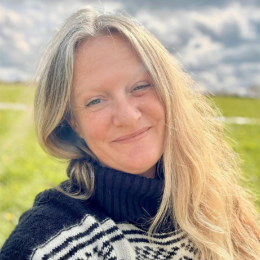 A woman with long blonde hair smiling outdoors on a cloudy day, wearing a black and white patterned sweater.