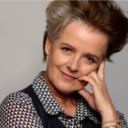 A confident middle-aged woman with short, gray hair, smiling gently while touching her face, wearing a black and white patterned blouse.