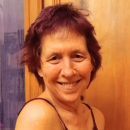 A woman with short, reddish-brown hair smiling at the camera, wearing a black spaghetti strap top, in front of a wooden background.