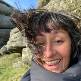 A woman smiling outdoors with rocky terrain and grass in the background.