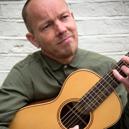 Man with short hair and a beard playing an acoustic guitar outdoors against a white brick wall, wearing a green shirt