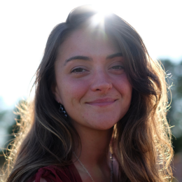 Young woman smiling outdoors with sunlight behind her, long wavy hair, and casual clothing.
