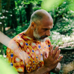 Man in colorful shirt with hands together in prayer or meditation outdoors in a lush green garden.