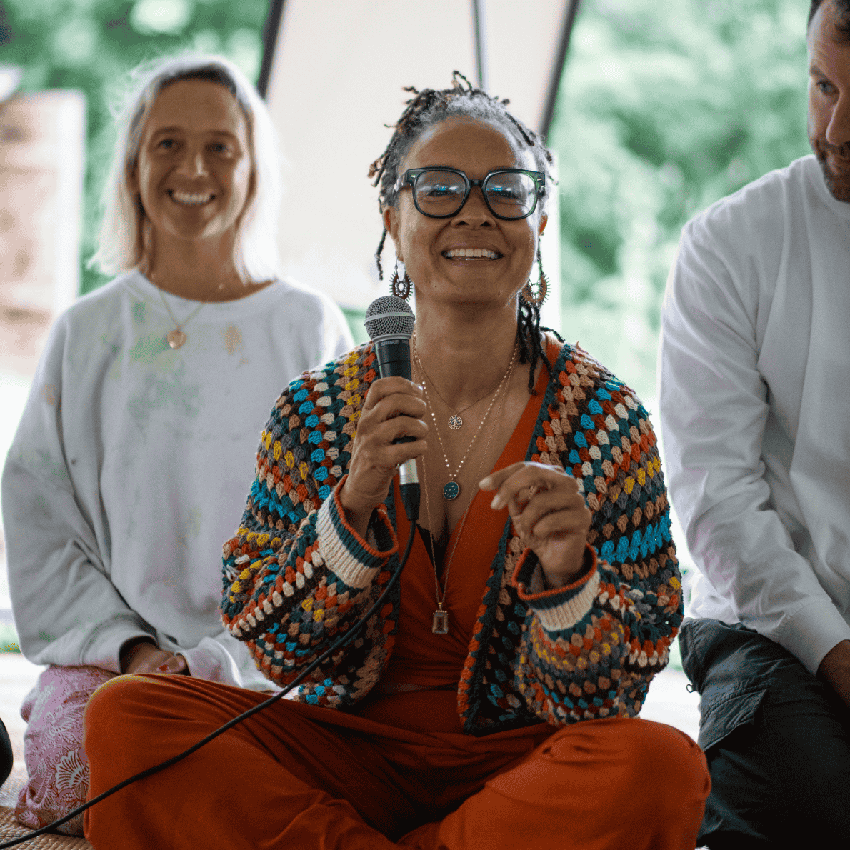 A woman with glasses and dreadlocks holding a microphone, sitting cross-legged in front of large windows with green trees outside, smiling while participating in an indoor gathering.