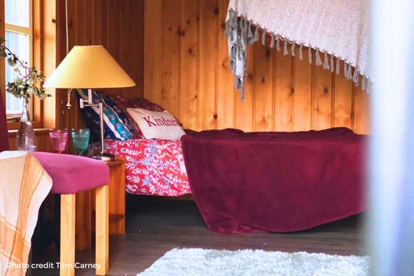 Cozy bedroom with wood-paneled wall, bed with red and white blankets, decorative pillows, a table lamp, and hanging tapestry or blanket on wall.