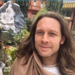 A man with long brown hair standing outdoors in front of a waterfall and green trees.