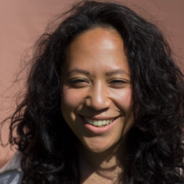 Close-up of a smiling woman with curly black hair, wearing a gray top, against a brown background.