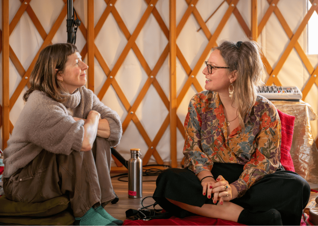 Two women sitting on the floor in a yurt, engaged in a conversation. One woman has short brown hair, is wearing a beige sweater, and has her arms crossed on her knees. The other woman has long wavy hair, glasses, and is wearing a colorful floral shirt. There is a water bottle, some electronic equipment, and a lamp in the background.