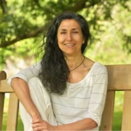 Smiling woman with long dark hair sitting on a wooden bench outdoors, surrounded by green trees.