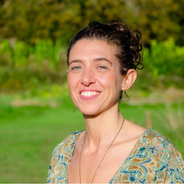 A young woman with dark curly hair smiling outdoors, wearing a patterned top, with greenery and trees in the background.