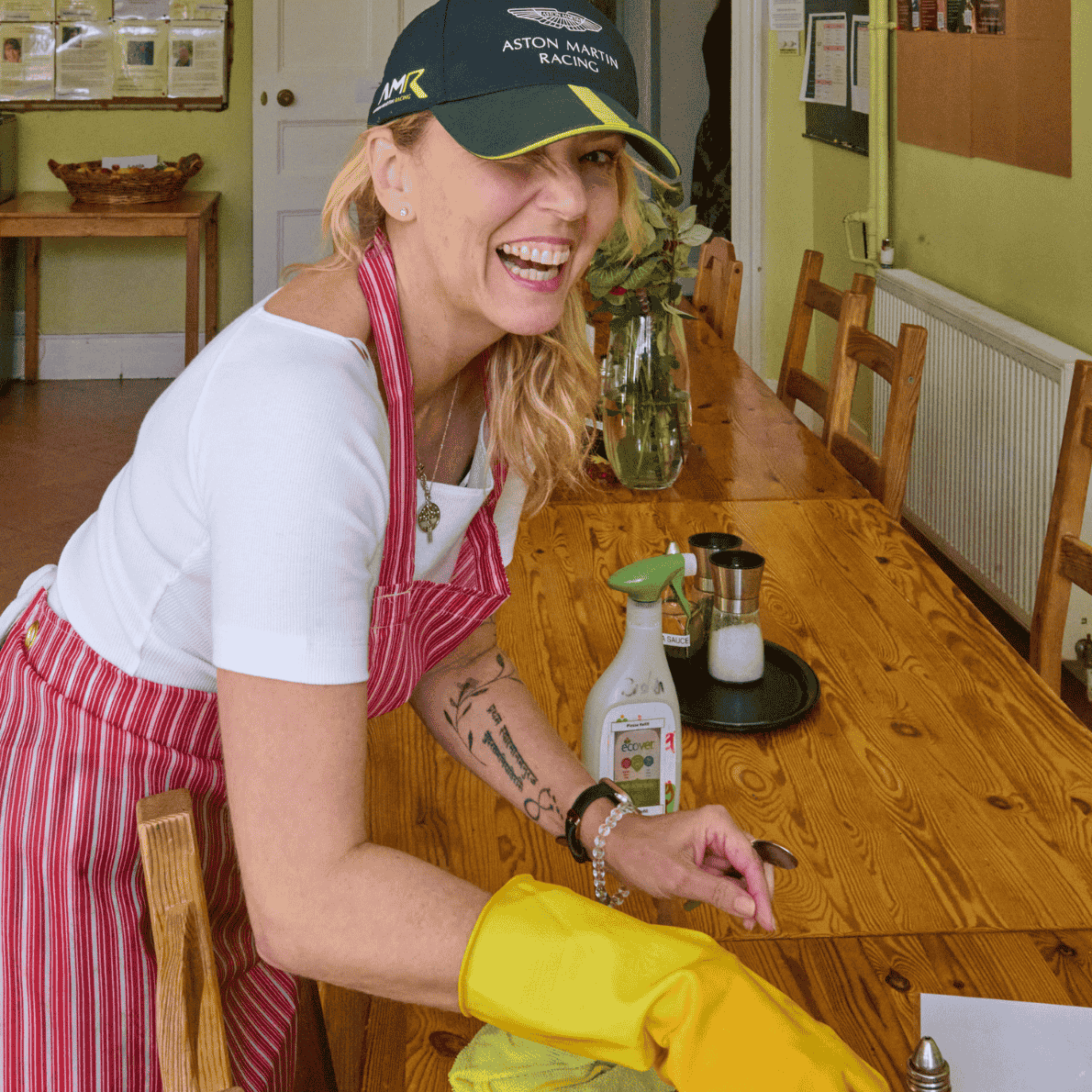 A woman in a striped apron and yellow rubber gloves cleaning a wooden dining table in a kitchen or dining area, smiling and looking at the camera.