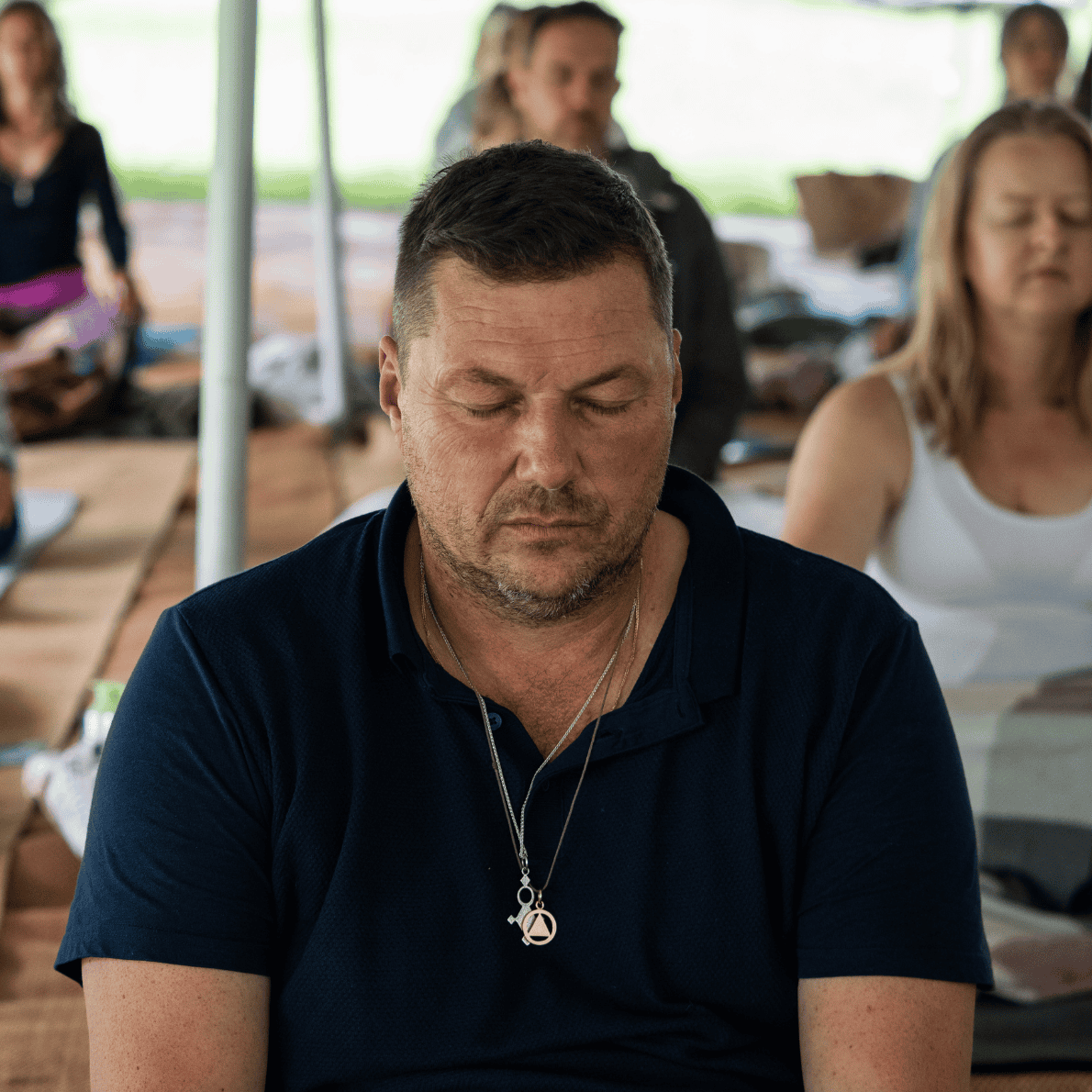 Man with closed eyes sitting with people in the background, under a canopy.