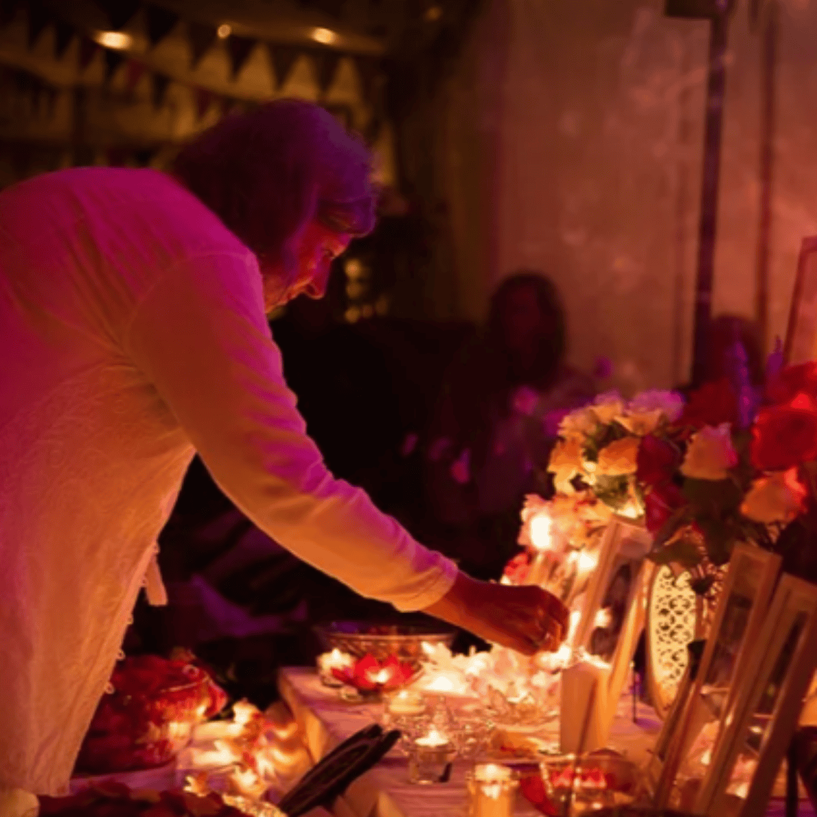 A woman lighting a candle at a memorial with flowers, photographs, and candles inside a dimly lit room, likely during a remembrance or tribute ceremony.