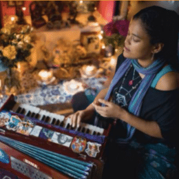 A young woman sitting at a decorated piano, looking at her phone, with a Christmas tree and holiday decorations in the background.