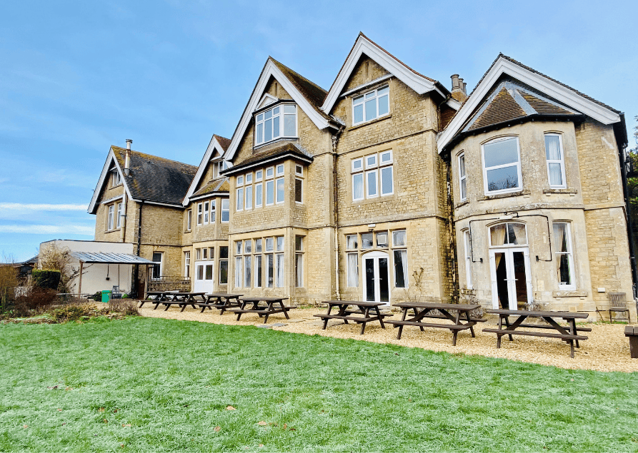 A large historic stone building with multiple windows and gabled roofs, surrounded by a lawn and picnic tables outside.