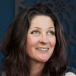 A woman with long brown hair smiling, wearing a dark top, with a decorative blue background.