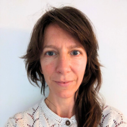 A woman with shoulder-length brown hair, wearing a white, crocheted top, standing in front of a plain, light-colored background.