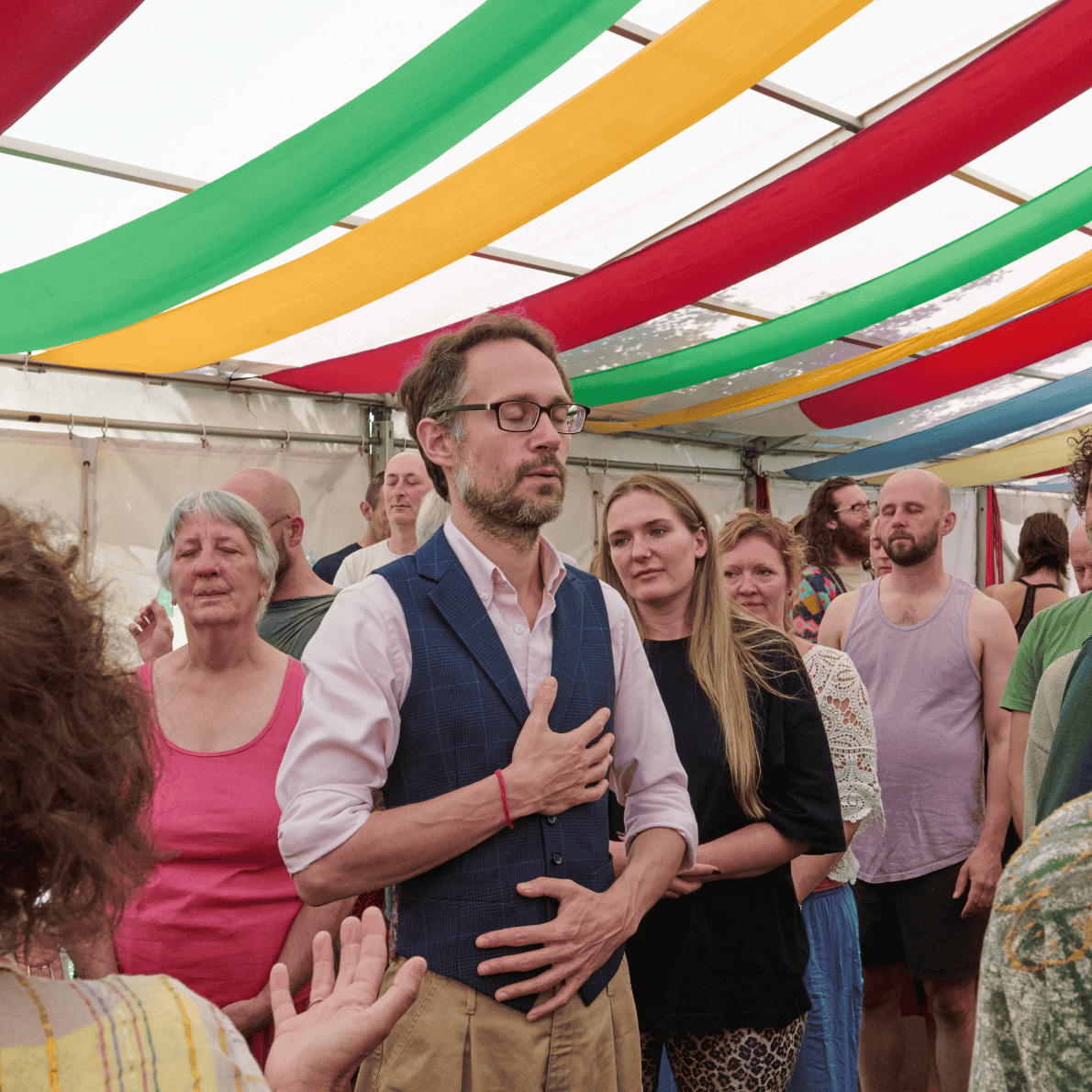People standing in a group inside a colorful tent, with some members holding their hands over their hearts and eyes closed, engaging in a moment of reflection or prayer.