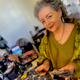 An older woman with gray curly hair wearing a green blouse, smiling while looking at her cards in a room filled with various items.