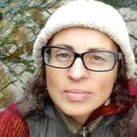 A woman with glasses wearing a white knit hat outdoors, with blurred trees and snow in the background.