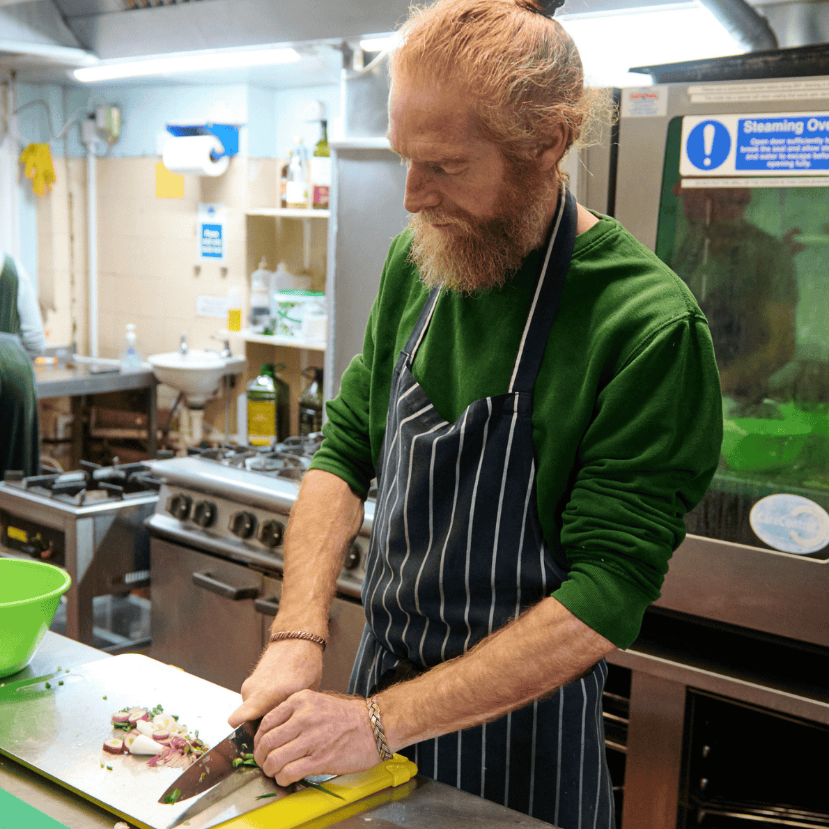 A man with a red beard and green sweater chopping vegetables on a yellow cutting board in a kitchen, wearing a black and white striped apron.