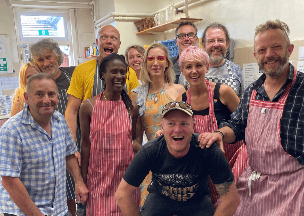 Group of people in a kitchen, smiling and posing for the camera, some wearing aprons.
