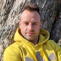 A man with short dark hair and a beard wearing a yellow jacket, standing outdoors in front of a large tree trunk.