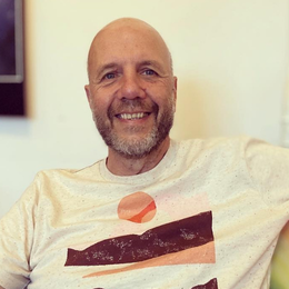 Smiling man with a beard and shaved head wearing a graphic T-shirt in an indoor setting.