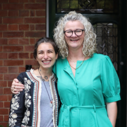 Two women smiling and standing close together outside a building with a brick wall and window behind them.