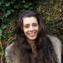 A woman with long dark brown hair, smiling, outdoors in front of green foliage and moss-covered rocks.