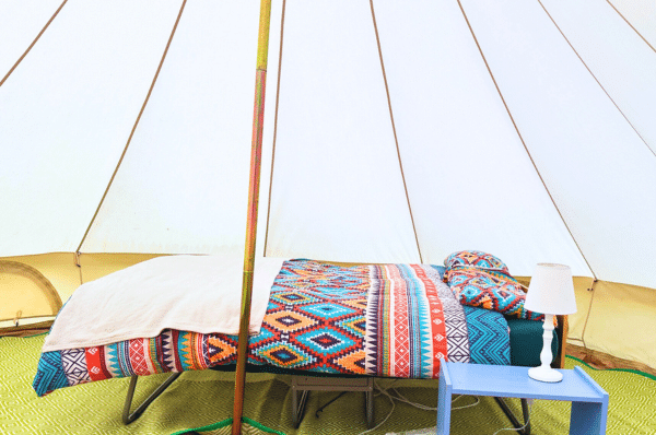 Interior of a canvas tent with a bed covered in a colorful, patterned blanket, a white pillow, a small blue table with a white lamp, and a green carpet.