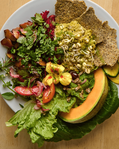 A white plate filled with mixed greens, sliced cucumbers, a slice of cantaloupe, sprouts, chopped vegetables, fresh herbs, and three pieces of whole grain bread, with some creamy spread or pasta in the middle.