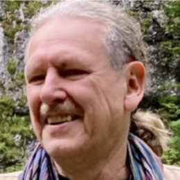 Close-up of a man smiling outdoors with grey hair and a plaid shirt, with trees and greenery in the background.