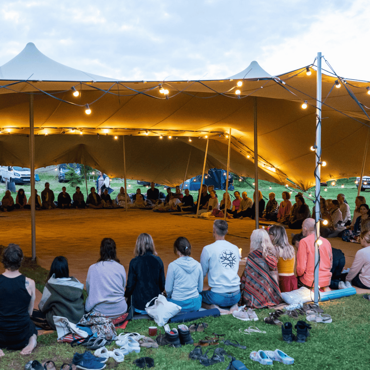 People sitting on the ground and under a large tent with string lights, attending an outdoor event in the evening.