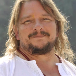 A man with long hair, a beard, and mustache smiling outdoors, wearing a white shirt.
