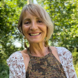 A smiling woman with short blonde hair outdoors, wearing a lace dress and necklace, surrounded by green trees.