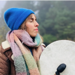 Woman wearing a blue knit hat and multicolored scarf, holding a large drum outdoors in a foggy, wooded area.