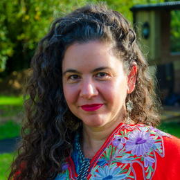 A woman with long, curly hair and a red lipstick, wearing a colorful, floral-patterned top, standing outdoors against a green background with trees and a structure.