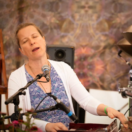 Woman singing into microphone, playing piano in a room with a stone wall background.