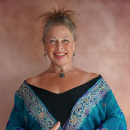 Smiling woman wearing a colorful shawl and jewelry, standing against a plain background.