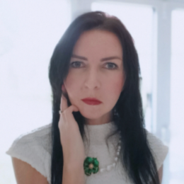 A woman with long dark hair, wearing a white top and a green floral necklace, posing with her hand near her face indoors.